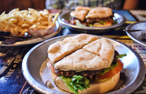 Two burgers with lettuce, tomato, and pickles on metal plates, accompanied by a side of French fries.