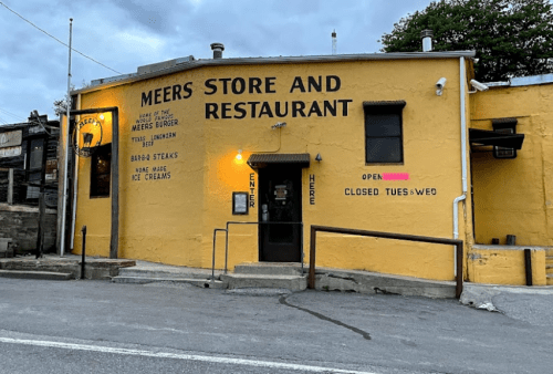 A yellow building with "Meers Store and Restaurant" signage, featuring a door and windows, set against a cloudy sky.