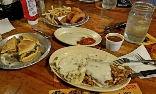 A plate with biscuits and gravy, a burger, fries, onion rings, and a cup of sauce on a wooden table.