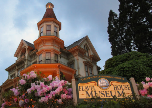 Historic Flavel House Museum with a Victorian-style architecture, surrounded by blooming flowers at dusk.