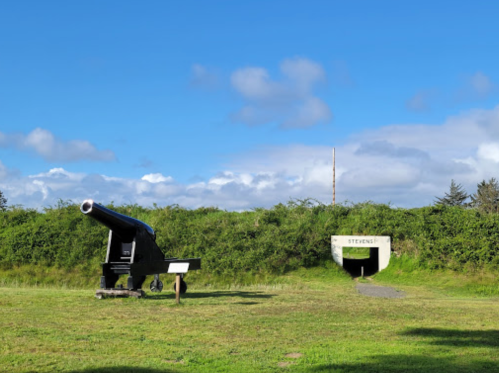 A historic cannon on a grassy field next to a bunker entrance, under a blue sky with scattered clouds.