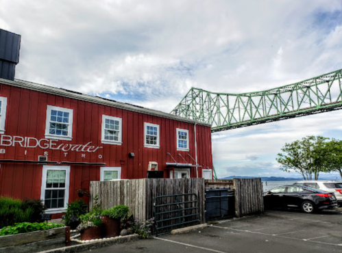 A red bistro building with a sign, next to a green bridge and a parking area by the water under a cloudy sky.