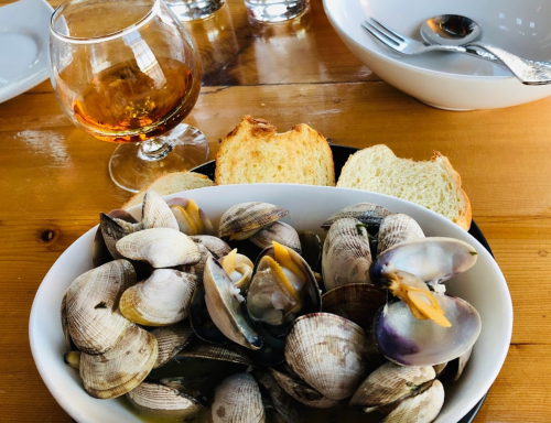 A bowl of clams served with slices of bread and a glass of amber liquid on a wooden table.