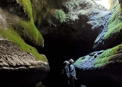 Two people stand in a moss-covered cave, illuminated by light from above, surrounded by rocky formations.