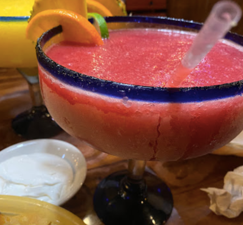 A vibrant frozen cocktail in a large glass, garnished with fruit slices, next to a bowl of sour cream.