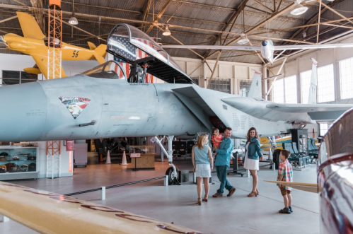 A family explores an aviation museum featuring a gray fighter jet and vintage aircraft in the background.