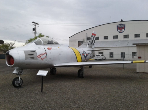 A silver military jet with yellow and black markings displayed outside a hangar.