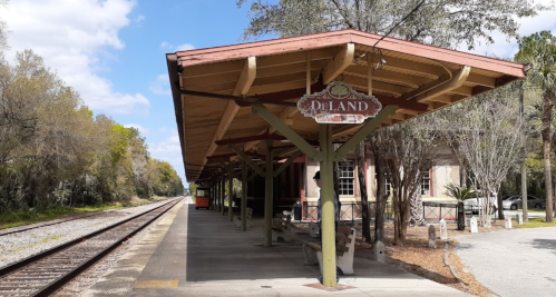 A train station platform with a sign reading "DeLand," surrounded by trees and a railway track.