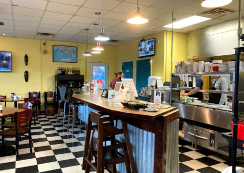 A cozy diner with a checkered floor, bar seating, and a visible kitchen area. Walls are decorated with maps and art.