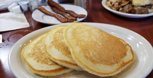 Three golden pancakes stacked on a white plate, with crispy bacon and a side of potatoes in the background.