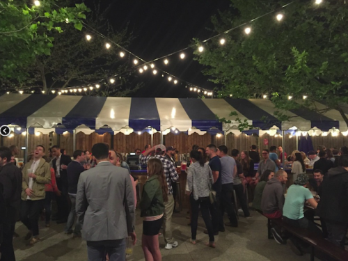 A lively outdoor gathering under string lights, with people socializing around tables and a tent in the background.