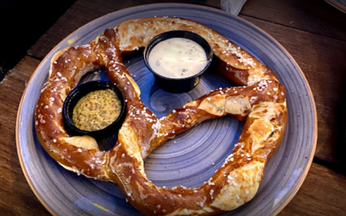 A large, soft pretzel on a plate, served with mustard and cheese dipping sauces.