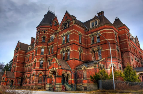 A large, historic brick building with intricate architecture, featuring multiple gables and a cloudy sky overhead.