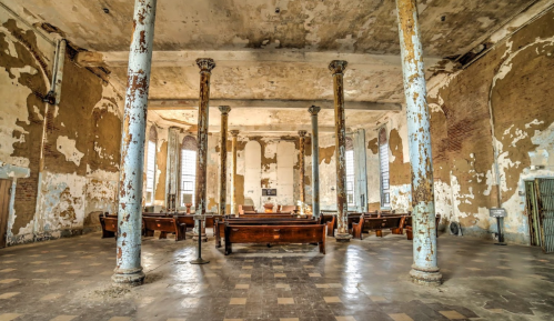 Interior of an abandoned building with peeling walls, tall columns, and wooden benches arranged in a circular layout.