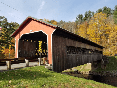 A red covered bridge spans a small stream, surrounded by autumn foliage and trees.