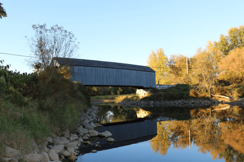 A covered bridge spans a calm river, reflecting autumn trees and a clear blue sky.