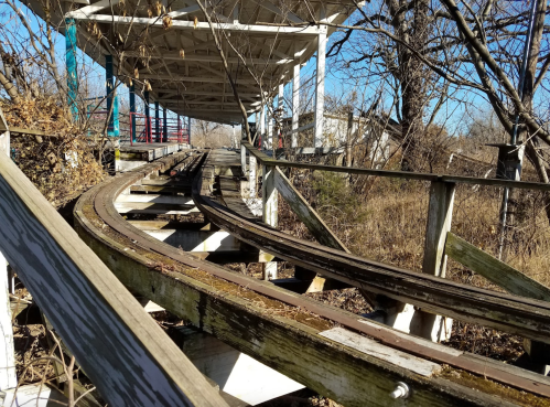 Abandoned roller coaster tracks winding through overgrown vegetation under a dilapidated structure.