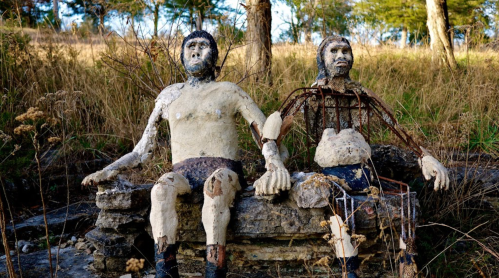Two abstract sculptures of figures sitting on a stone bench, surrounded by grass and trees.