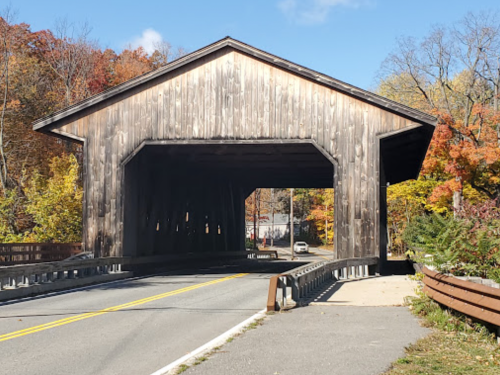 A wooden covered bridge spans a road, surrounded by autumn foliage and a clear blue sky.