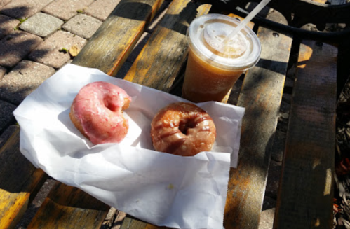 Two donuts on a white napkin and a cup of iced coffee on a wooden bench.