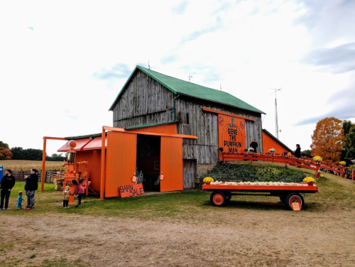 A rustic barn painted orange, with pumpkins and a vibrant garden, surrounded by visitors enjoying the autumn scenery.