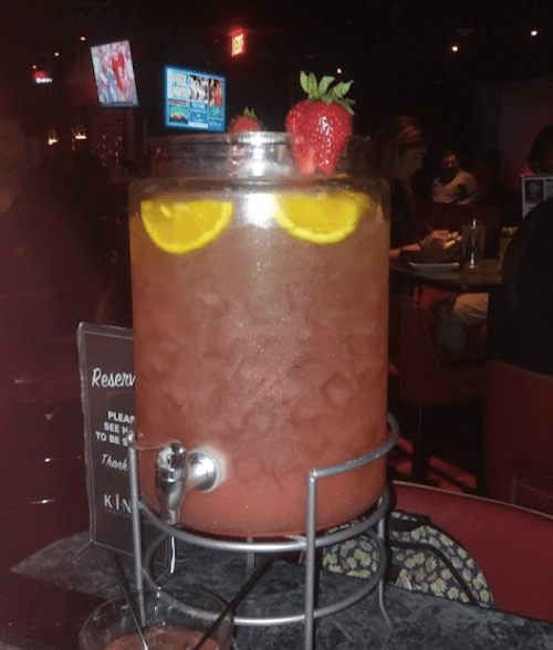 A large beverage dispenser filled with a fruity drink, garnished with lemon slices and a strawberry, in a dimly lit bar.