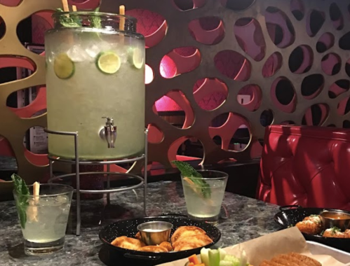 A beverage dispenser filled with lime-infused drink, surrounded by glasses and plates of appetizers on a stylish table.