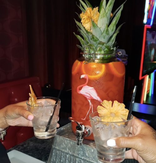 Two hands hold glasses of clear drinks in front of a large, pineapple-topped beverage dispenser with a flamingo design.