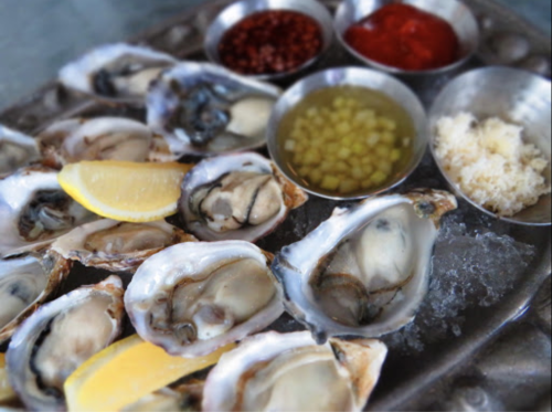 A platter of fresh oysters on ice, served with lemon wedges and various sauces in small bowls.