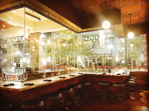 A modern restaurant interior with a bar, stools, and large windows showcasing an outdoor view. Warm lighting creates a cozy atmosphere.