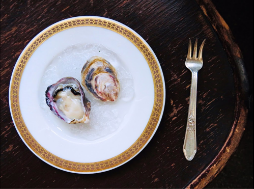 A plate with two oysters on ice, accompanied by a silver fork, set on a wooden table.