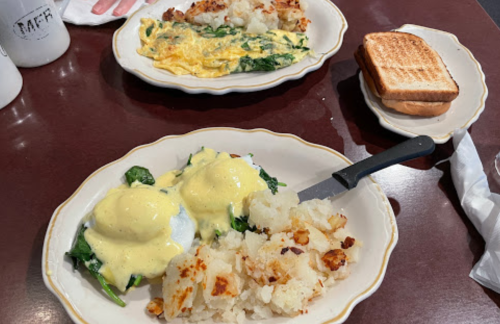 Two plates of breakfast: one with eggs Benedict on spinach and hash browns, the other with an omelet and toast.