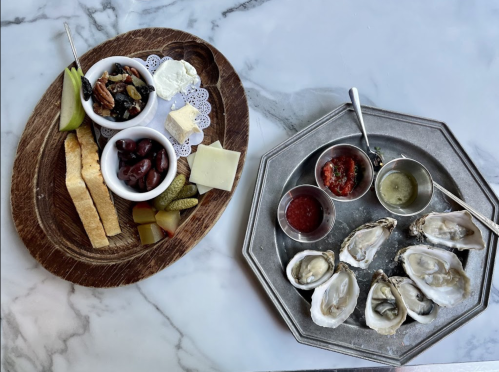 A wooden platter with olives, cheese, and crackers beside a metal tray of oysters and dipping sauces on a marble surface.