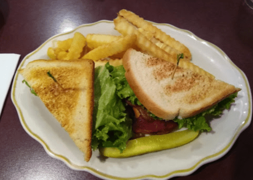 A plate with a toasted sandwich filled with lettuce and bacon, accompanied by crinkle-cut fries and a pickle.