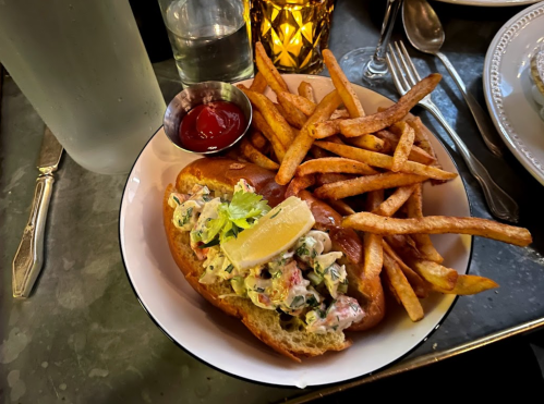 A plate with a lobster roll topped with lemon, served with fries and a small cup of ketchup.