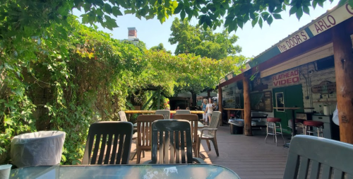 Outdoor seating area of a cafe, surrounded by greenery and rustic decor, with tables and chairs under a sunny sky.