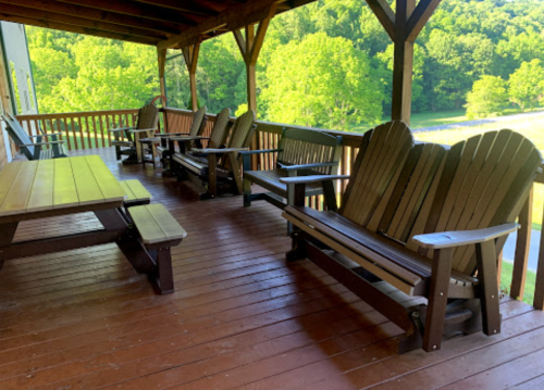 A wooden porch with several Adirondack chairs and a picnic table, surrounded by green trees.