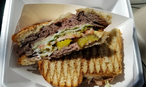 A close-up of a sliced sandwich with layers of roast beef, lettuce, tomato, pickles, and grilled bread.