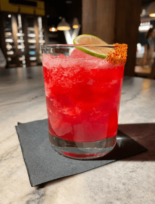 A vibrant red cocktail with a lime slice and a salted rim, served in a glass on a black napkin.