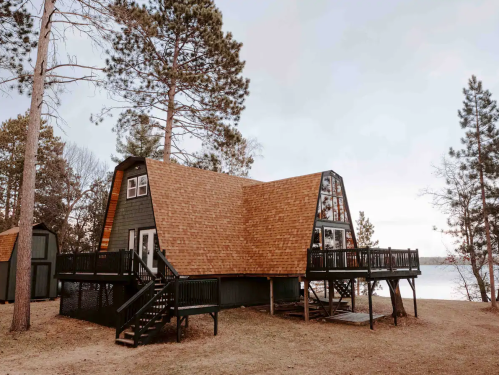 A modern A-frame cabin with a wooden deck, surrounded by tall trees near a lake.