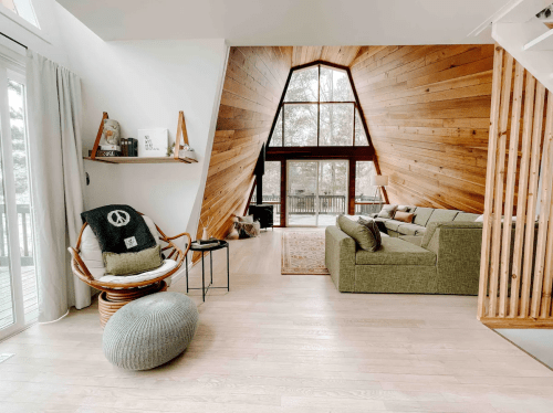 Cozy living room with wooden walls, large windows, a green sofa, and a stylish chair, creating a warm atmosphere.