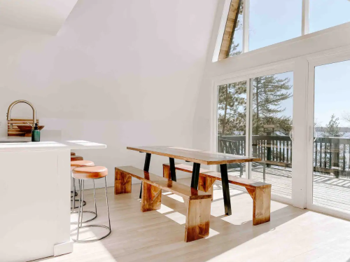 Bright, modern interior featuring a wooden dining table, bar stools, and large windows with a view of the outdoors.