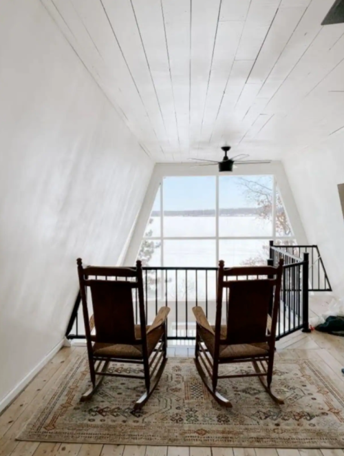 Two wooden rocking chairs facing a large window with a view of a lake, set in a bright, minimalist room.