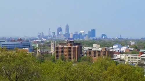 A panoramic view of a city skyline with tall buildings, greenery in the foreground, and a clear blue sky.