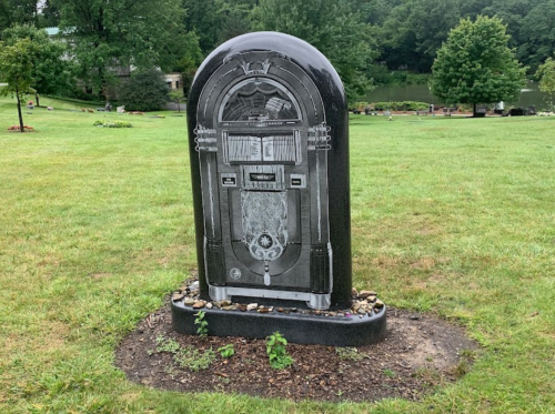 A black granite gravestone shaped like a vintage jukebox, surrounded by grass and small stones.
