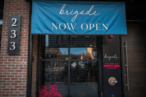 A storefront with a blue banner reading "Brigade NOW OPEN" and a pink logo on the door. Brick wall and address visible.