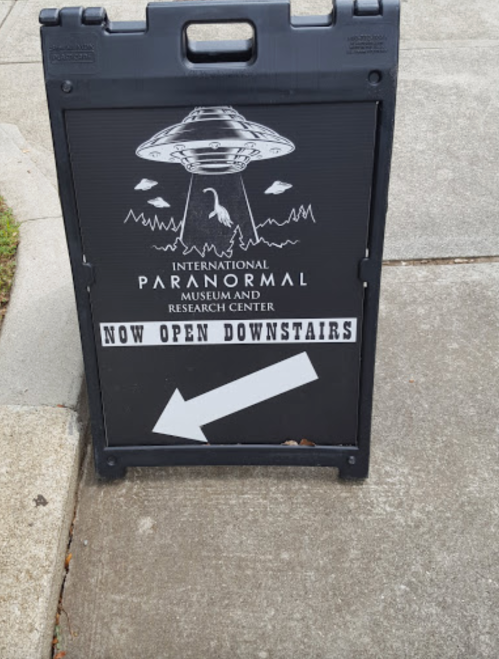 A sign for the International Paranormal Museum and Research Center, indicating it's now open downstairs, with a UFO graphic.