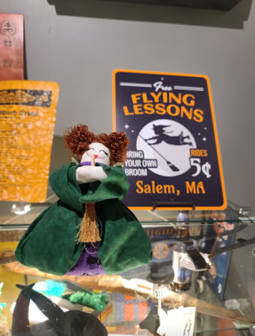 A whimsical witch doll in a green cloak stands in front of a sign for free flying lessons in Salem, MA.