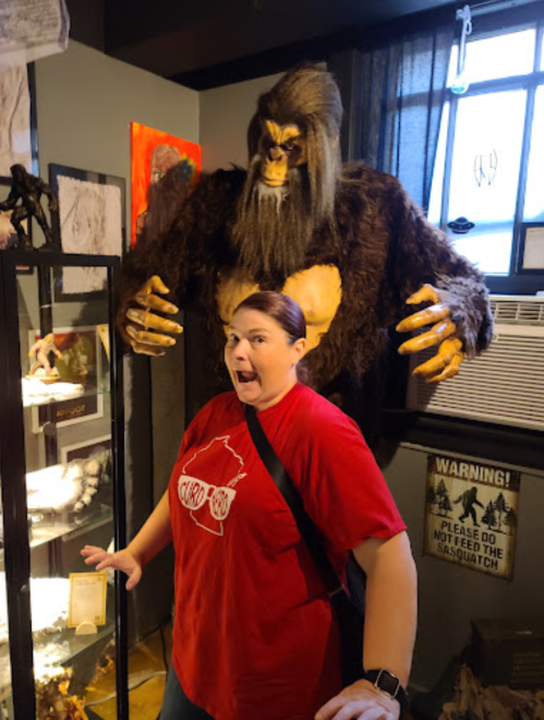 A woman in a red shirt poses playfully with a large Sasquatch statue in a museum setting.