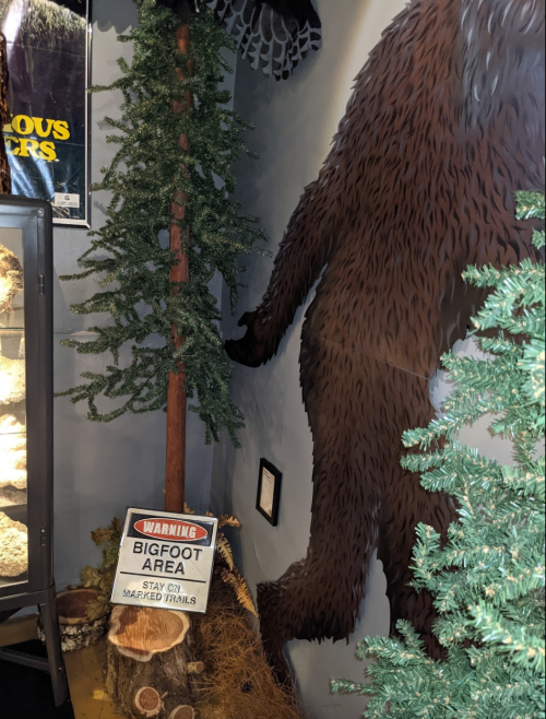 A large Bigfoot figure stands next to a tree, with a warning sign about Bigfoot nearby in a themed display.